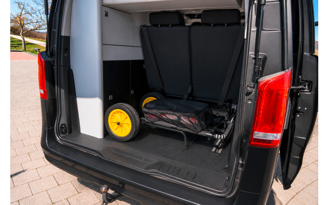 Geöffnete Hecktüren eines schwarzen Vans, Blick in den Laderaum; auf dem Boden liegt ein zusammengeklapptes schwarzes Gerät mit zwei gelben Rädern, Pflaster und Teil der Rückleuchte sichtbar