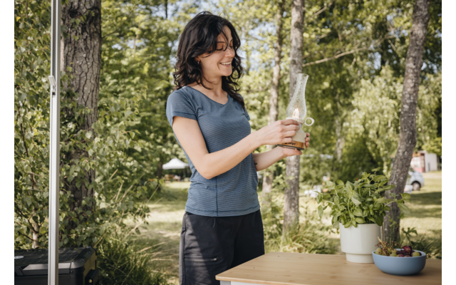 Frau steht draußen an einem Tisch und gießt Wasser aus einer Glasflasche in ein Glas; Töpfe mit Pflanzen und Bäume im Hintergrund