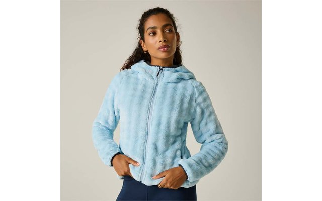 Persona con el cabello oscuro recogido lleva una chaqueta con capucha y cremallera de tejido peludo azul claro, manos en los bolsillos, leggings oscuros, de pie frente a un fondo neutro y claro, mirando ligeramente de lado
