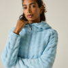 Retrato de medio cuerpo de una mujer con el cabello oscuro recogido, que lleva una chaqueta con capucha de pelo sintético azul claro con cremallera y sujeta el cuello con una mano; fondo liso y claro