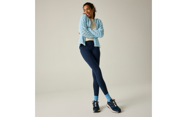 Persona de pie con las piernas cruzadas, brazos cruzados sobre el pecho, viste una chaqueta de forro polar azul claro, una camiseta blanca, mallas azul oscuro, calcetines azul claro y zapatillas deportivas azul oscuro sobre un fondo liso y neutro