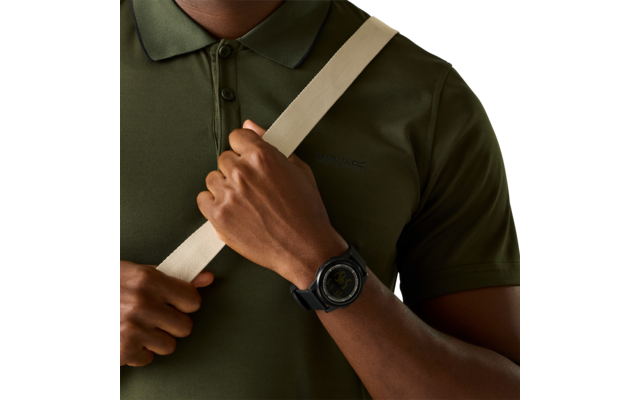 Bei­ge­schnittener Oberkörper einer Person in olivgrünem Poloshirt, ein beiger Riemen verläuft diagonal über die Brust, eine Hand hält den Riemen; am Handgelenk eine schwarze, runde Smartwatch