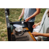 Two people at a portable camping kitchen on grass. One person holds a spoon over a pot and an open plastic container with white contents. On the countertop are a lidded pot, stacked plates, a sink and a dish rack