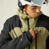 Person mit weißem Helm und hochgeschobener Skibrille trägt olivgrün-schwarze Jacke und zieht an einem neongrünen Reißverschluss-Anhänger