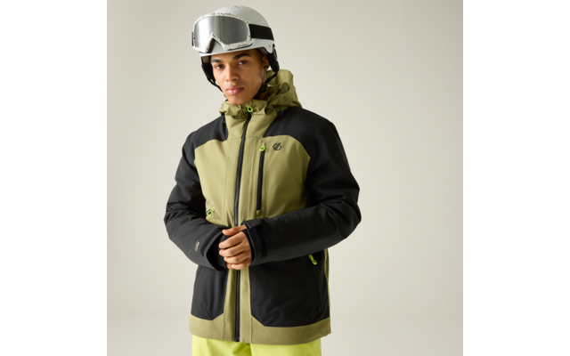 Person steht vor neutralem Hintergrund, trägt weißen Skihelm mit Skibrille, olivgrüne und schwarze Skijacke mit Kapuze und neonfarbigen Reißverschlüssen, hellgrüne Skihose, hält den Reißverschluss der Jacke mit beiden Händen