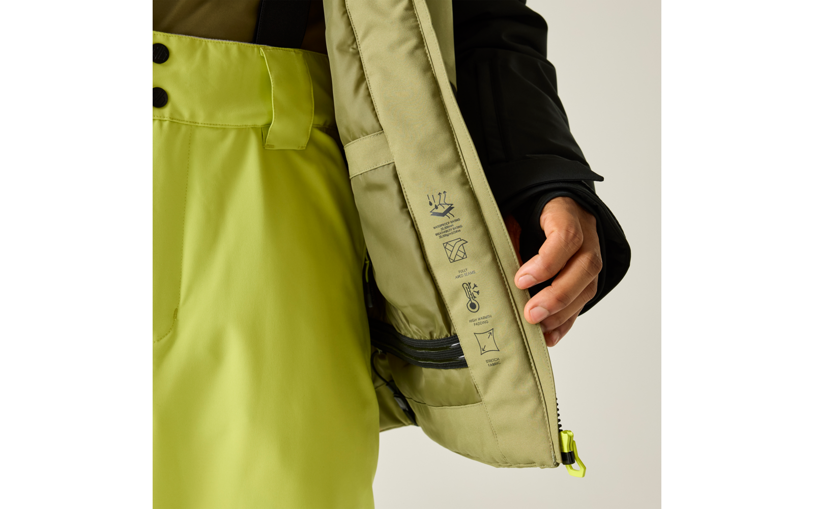 Hand hält eine geöffnete hellgrüne Jacke, Innenseite mit Pflegezeichen und Reißverschlussfach; hellgrüne Hose und schwarzer Jackenärmel sichtbar