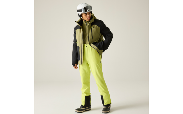 Person mit weißem Skihelm und Skibrille, olivegrüne und schwarze Jacke, leuchtend gelbgrüne Hose, schwarze Schneestiefel, steht auf hellem Hintergrund, Blick nach unten