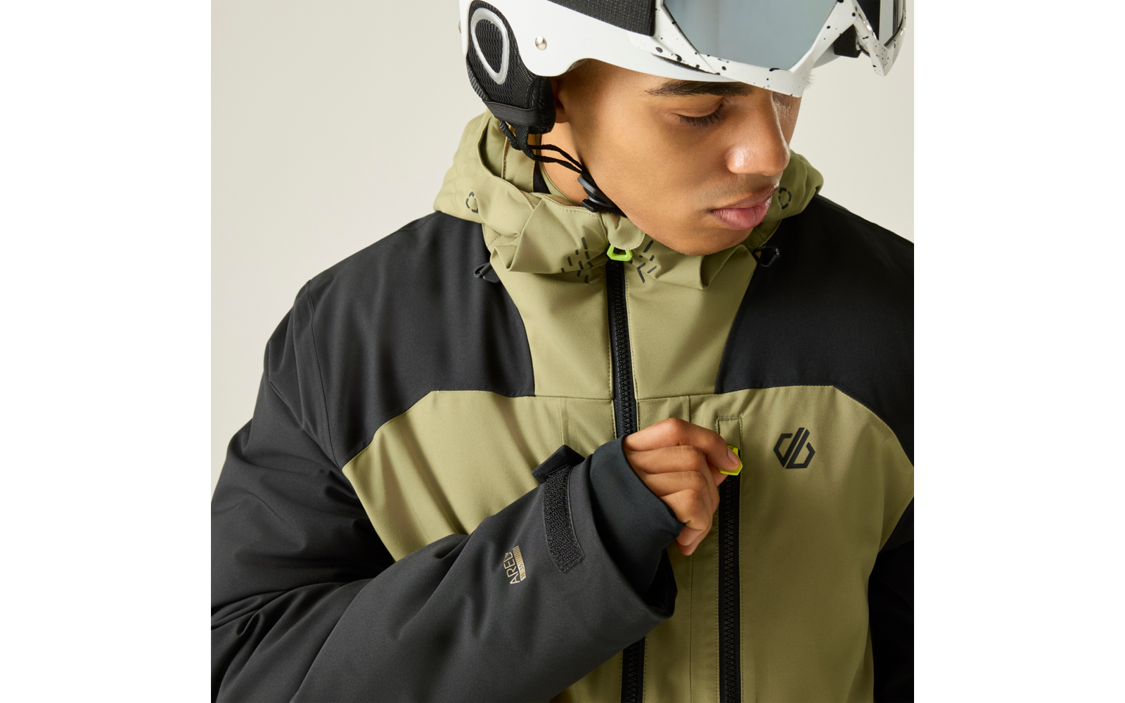 Person mit weißem Helm und hochgeschobener Skibrille trägt olivgrün-schwarze Jacke und zieht an einem neongrünen Reißverschluss-Anhänger