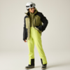 Person mit weißem Skihelm und Skibrille, olivegrüne und schwarze Jacke, leuchtend gelbgrüne Hose, schwarze Schneestiefel, steht auf hellem Hintergrund, Blick nach unten