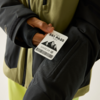Hand hält eine Karte mit der Aufschrift "SKI PASS", Bergsymbol und Barcode vor einer schwarzen und olivgrünen Jacke