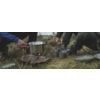 Topf auf kleinem Kocher auf dem Boden, Hände und Beine von Personen in Outdoor-Kleidung, mehrere Metallbecher und Teller auf Gras