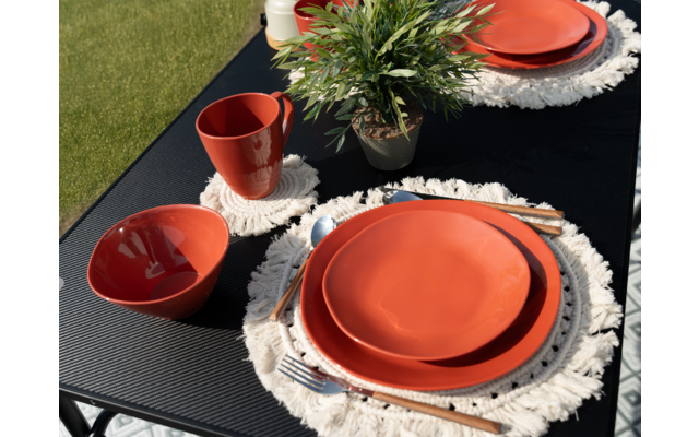 Ein gedeckter Tisch mit orangenen Tellern, Tassen und weißen Häkeluntersetzern auf einer schwarzen Tischdecke, im Hintergrund grünes Gras