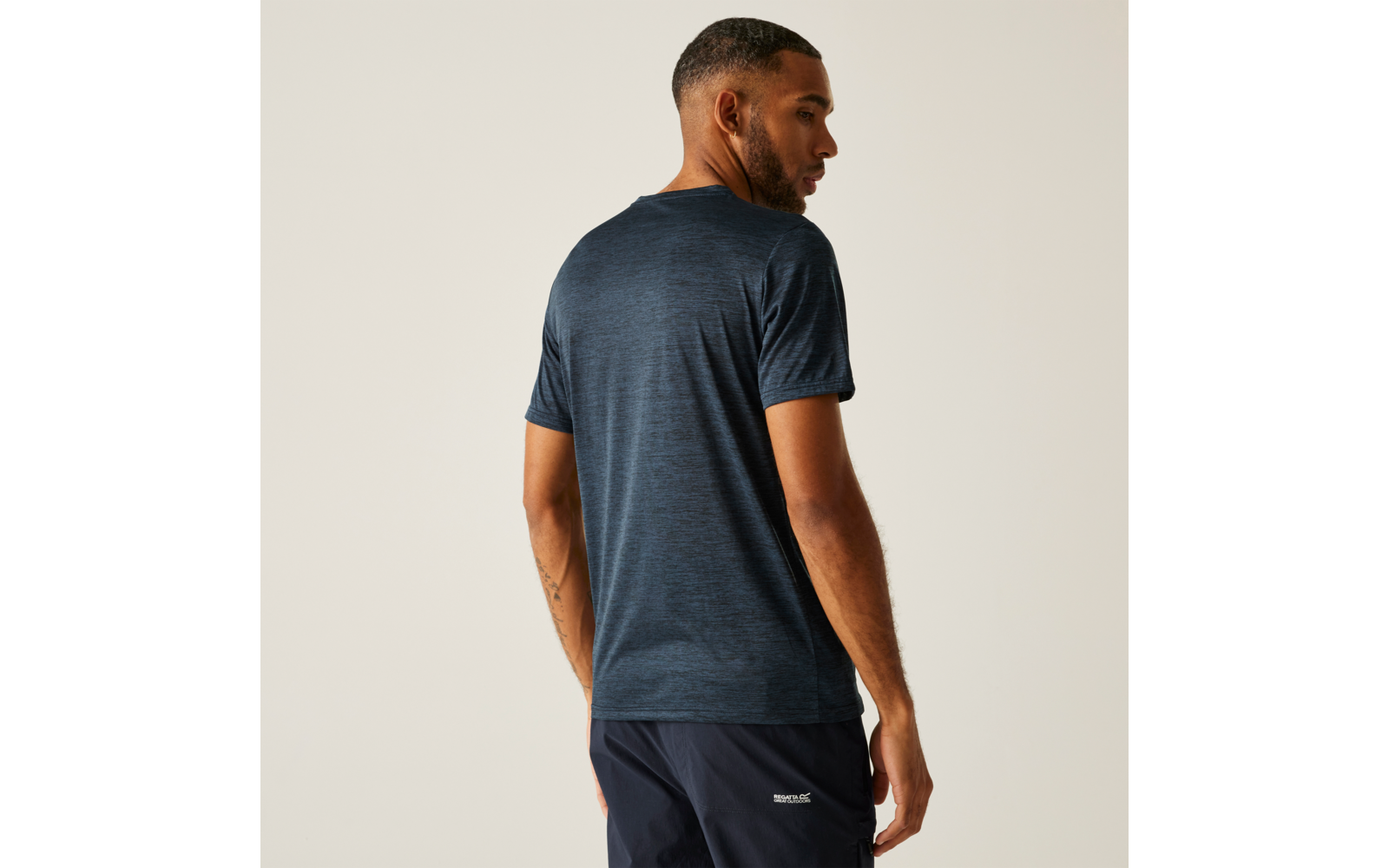 Person von hinten, trägt blaues kurzärmeliges T‑Shirt und dunkle Shorts mit kleinem weißem Logo, schaut über die Schulter, heller Hintergrund