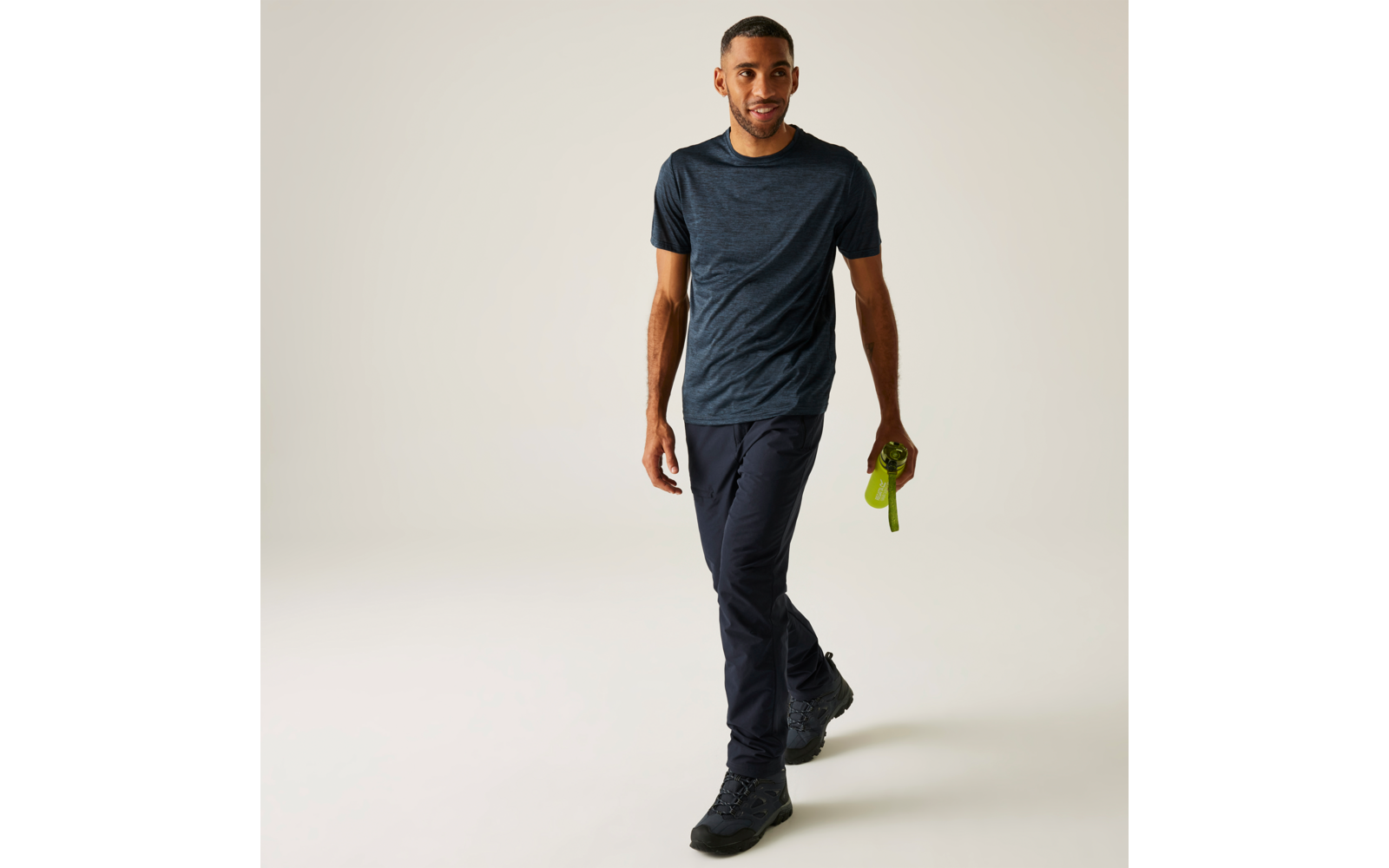 Mann in Freizeitkleidung geht auf hellem Hintergrund, trägt dunkelblaues T‑Shirt, dunkle Hose und schwarze Schuhe, hält eine grüne Trinkflasche in der rechten Hand