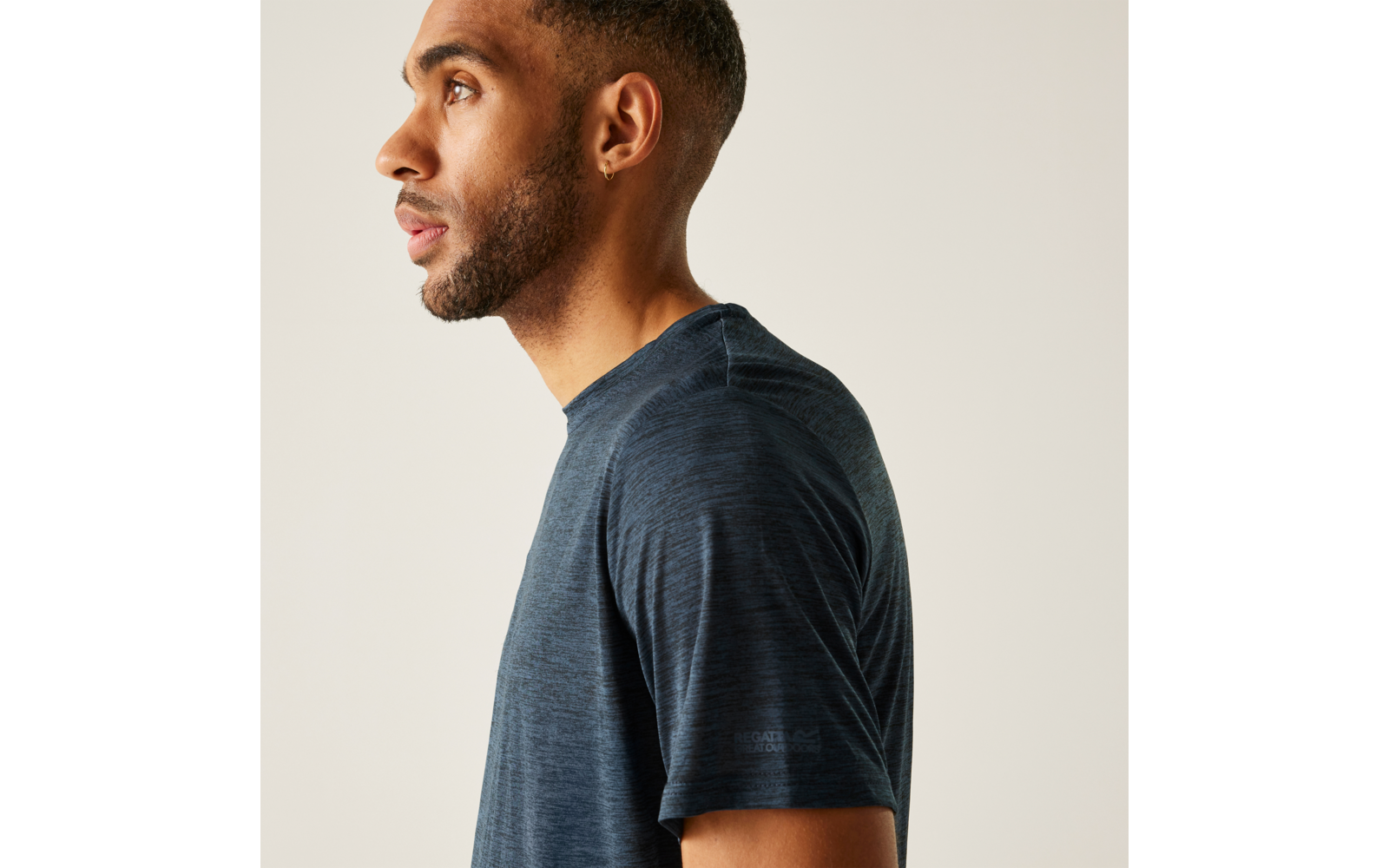 Seitliches Porträt einer Person mit kurzem Haar, Bart und Ohrstecker, trägt dunkelblaues T‑Shirt vor hellem Hintergrund