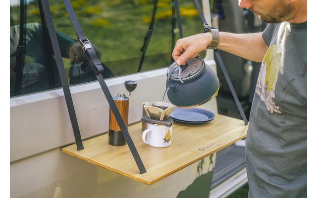 Person gießt Flüssigkeit aus einer Kanne durch einen Papierfilter in eine Tasse auf einem hölzernen Ausklapptisch; daneben stehen ein zylindrisches Metallgerät und eine Untertasse; der Tisch ist an der Außenseite eines Fahrzeugs befestigt