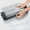 Twee handen voeren een transparante plastic zak door een zwart sluitingsapparaat
