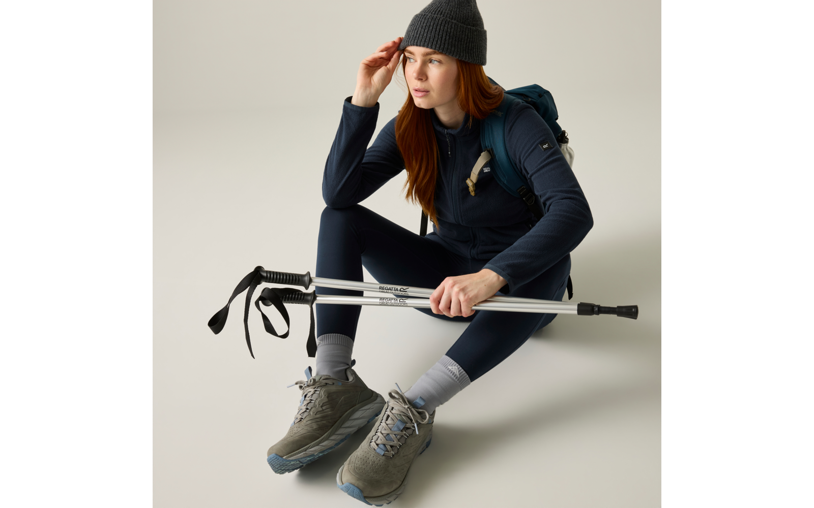 Persona con capelli rossi seduta a terra, indossa un berretto grigio, una giacca blu scuro e uno zaino, tiene due bastoncini da trekking ripiegati; indossa scarpe da trekking grigie e calzini grigio chiaro, guarda verso destra
