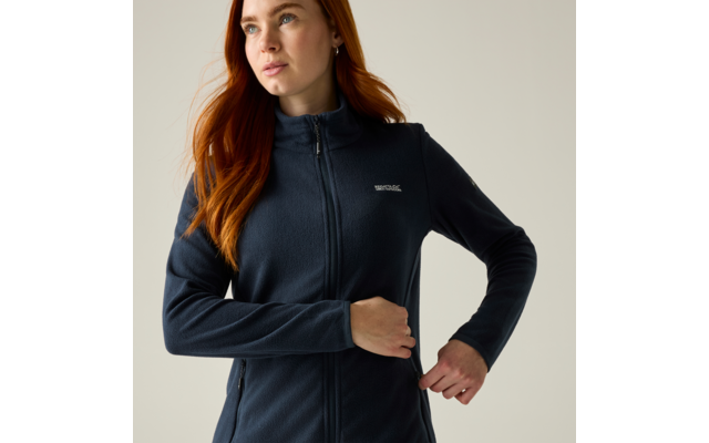 Persona con capelli rossi che indossa una giacca in pile blu scuro con cerniera, guarda in alto a sinistra, una mano vicino alla cerniera, sfondo chiaro uniforme