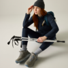 Persona con capelli rossi seduta a terra, indossa un berretto grigio, una giacca blu scuro e uno zaino, tiene due bastoncini da trekking ripiegati; indossa scarpe da trekking grigie e calzini grigio chiaro, guarda verso destra