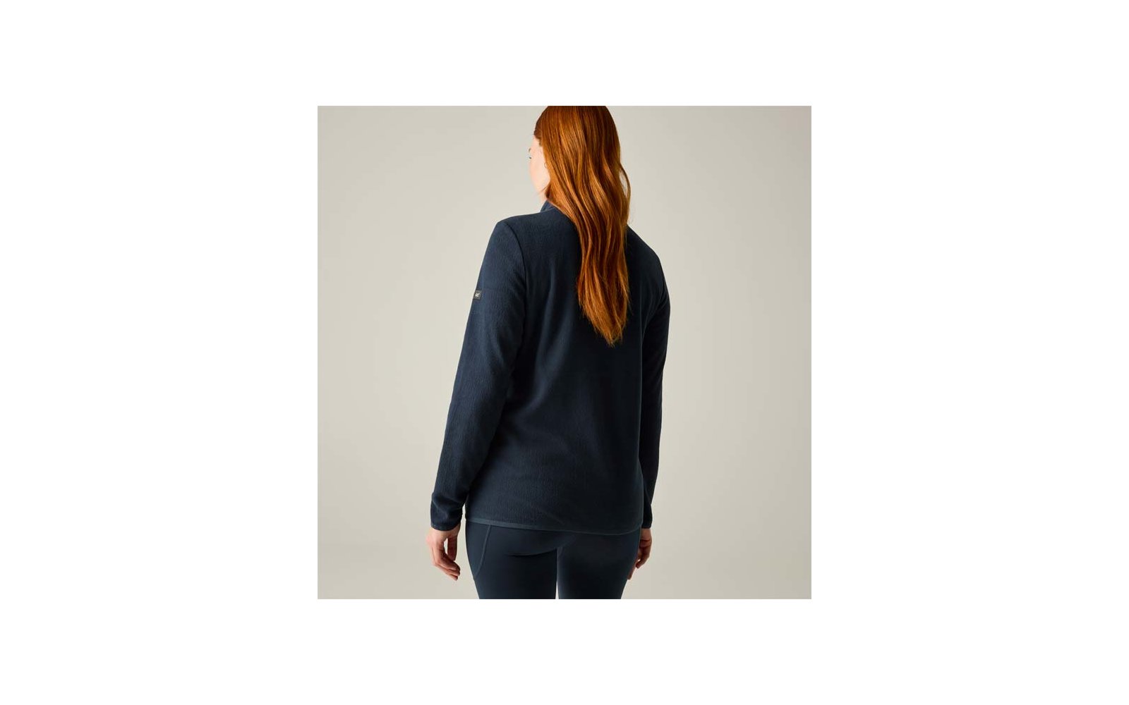 Vista trasera de una persona con cabello largo y rojo, viste una camiseta de manga larga azul marino y leggings, de pie sobre un fondo neutro claro