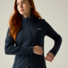 Persona con capelli rossi che indossa una giacca in pile blu scuro con cerniera, guarda in alto a sinistra, una mano vicino alla cerniera, sfondo chiaro uniforme