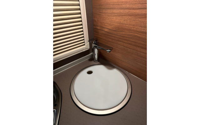 Ronde witte afdekking met metalen rand op een ronde opening, ééngreepskraan erboven, houten paneel op de achtergrond, raam met gesloten jaloezie links, grijs aanrecht