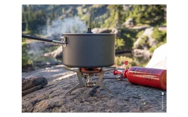 Topf mit Deckel auf kleinem tragbarem Kocher auf einem Felsen, Dampf steigt auf; rote Gasflasche daneben; See und Bäume im Hintergrund