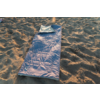 Sac de couchage bleu déployé avec partie supérieure repliée sur sol sablonneux, lumière du soleil et empreintes de pas dans le sable