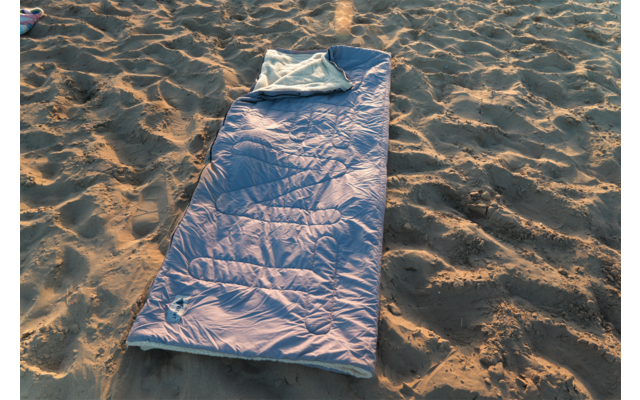 Sac de couchage bleu déployé avec partie supérieure repliée sur sol sablonneux, lumière du soleil et empreintes de pas dans le sable