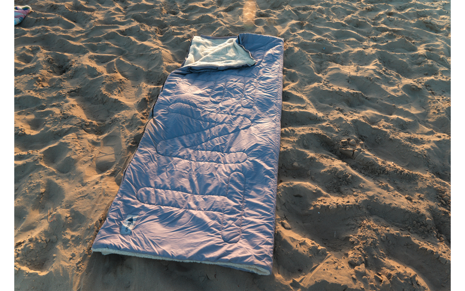 Sac de couchage bleu déployé avec partie supérieure repliée sur sol sablonneux, lumière du soleil et empreintes de pas dans le sable