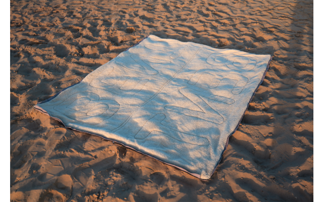 Couverture rectangulaire bleu clair posée sur du sable, ombres de feuilles de palmier sur la couverture
