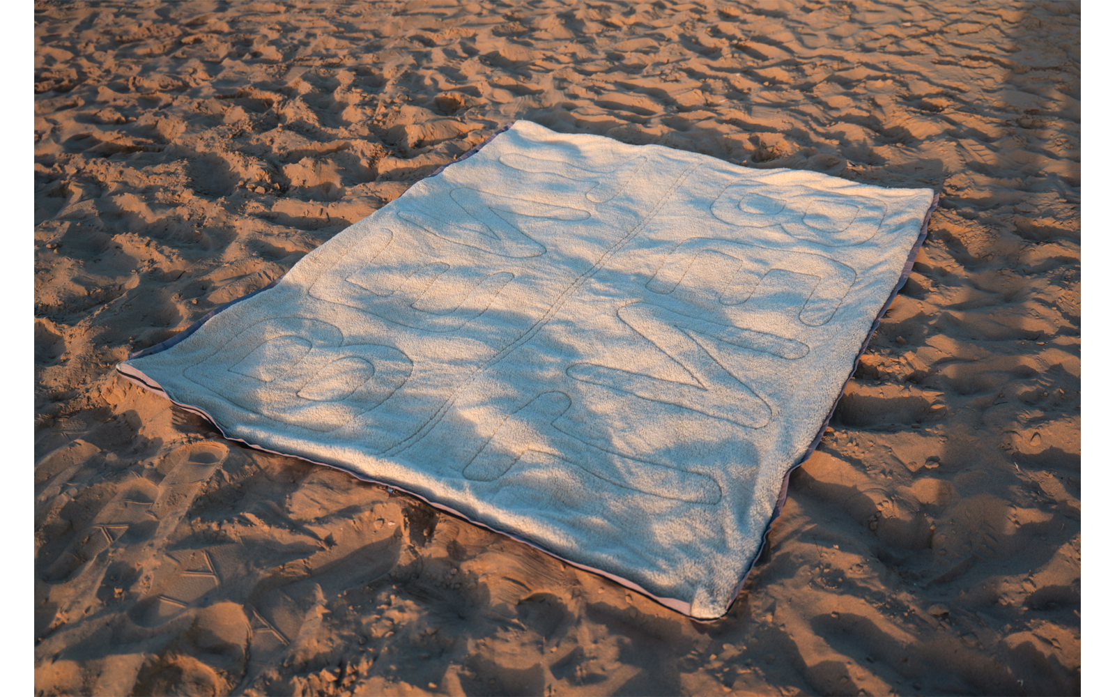 Couverture rectangulaire bleu clair posée sur du sable, ombres de feuilles de palmier sur la couverture