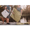 Deux personnes se tiennent sur un rocher au bord d’un lac en tenant une couverture matelassée verte ou un sac de couchage; un chien se tient à leur gauche; un sac vert et des vêtements sont posés sur le rocher; eau et arbres en arrière-plan