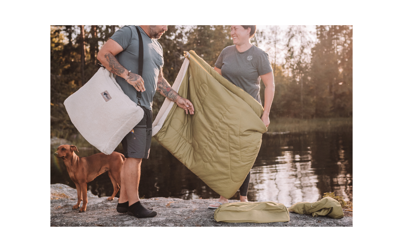 Deux personnes se tiennent sur un rocher au bord d’un lac en tenant une couverture matelassée verte ou un sac de couchage; un chien se tient à leur gauche; un sac vert et des vêtements sont posés sur le rocher; eau et arbres en arrière-plan
