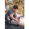Personne à genoux sur un rocher près de pins, portant un t‑shirt gris et un pantalon sombre, touchant un textile à motifs à côté d'un sac à dos