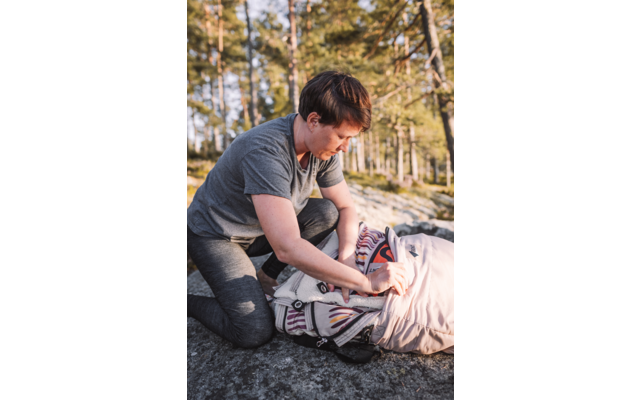 Personne à genoux sur un rocher près de pins, portant un t‑shirt gris et un pantalon sombre, touchant un textile à motifs à côté d'un sac à dos