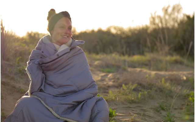 Personne portant un bandeau enveloppée dans un sac de couchage gris assise sur des dunes de sable avec des herbes, lumière du soleil en arrière-plan