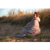 Personne assise sur une dune de sable, enveloppée dans une couette gris clair, regardant la caméra et souriant, herbes de dunes en arrière‑plan