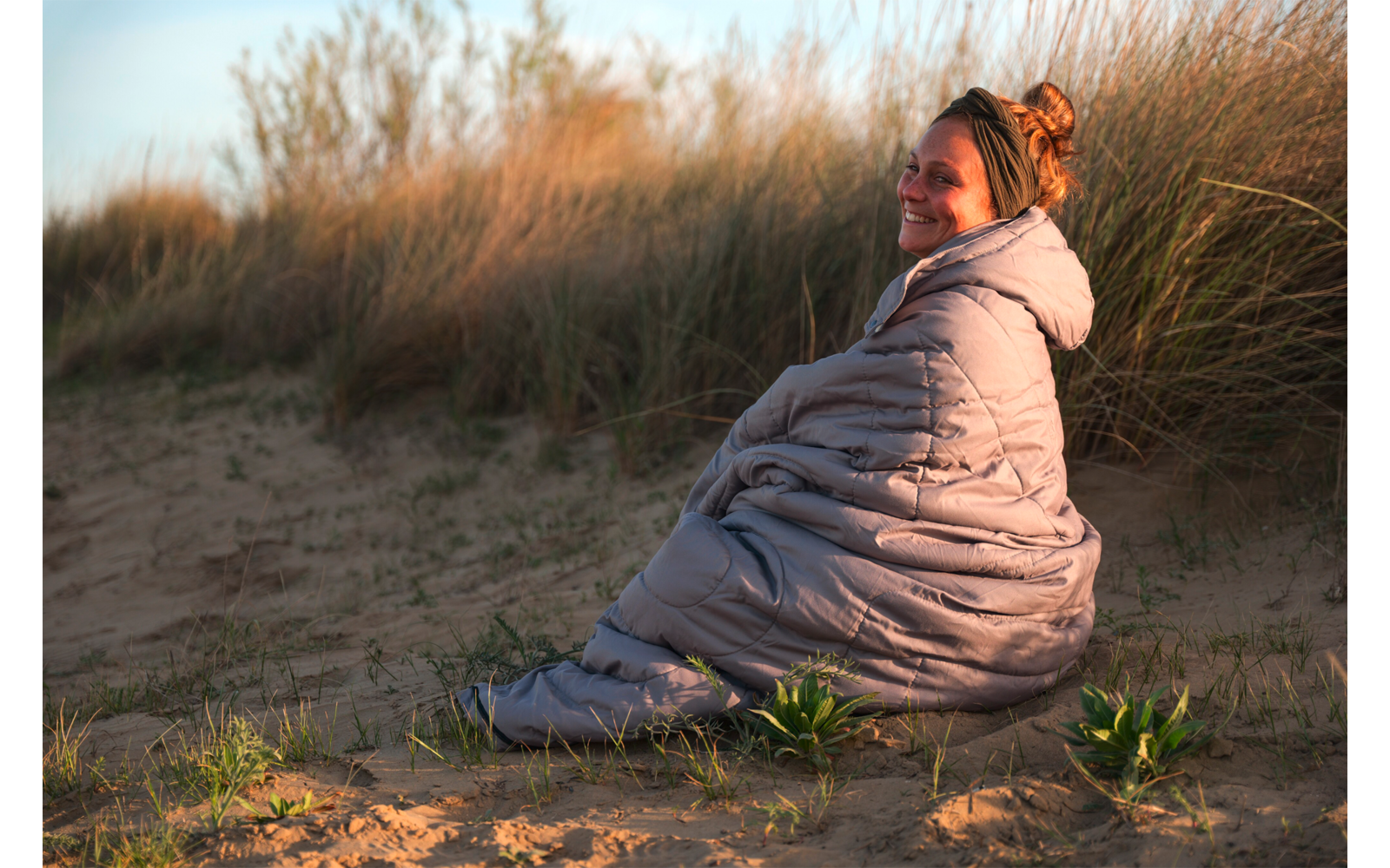 Personne assise sur une dune de sable, enveloppée dans une couette gris clair, regardant la caméra et souriant, herbes de dunes en arrière‑plan