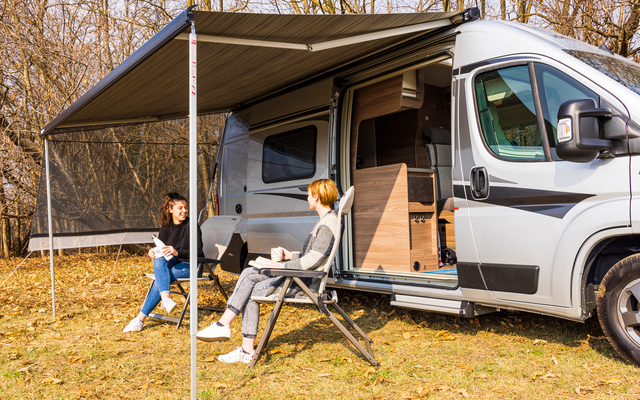 Wohnmobil mit ausgefahrener Markise; zwei Personen sitzen auf Campingstühlen vor dem offenen Seiteneingang; Wiese mit kahlen Bäumen im Hintergrund