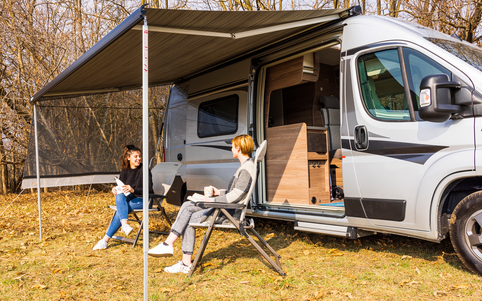 Wohnmobil mit ausgefahrener Markise; zwei Personen sitzen auf Campingstühlen vor dem offenen Seiteneingang; Wiese mit kahlen Bäumen im Hintergrund