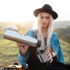 Woman with blonde hair, black hat and denim jacket pours liquid from a thermos into a metal cup; she sits on grass, a car and hills are visible in the background