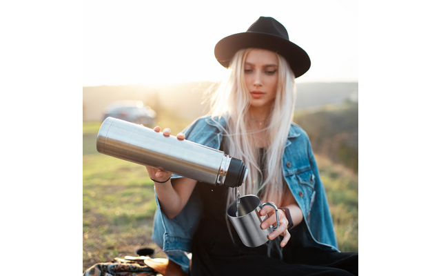 Woman with blonde hair, black hat and denim jacket pours liquid from a thermos into a metal cup; she sits on grass, a car and hills are visible in the background