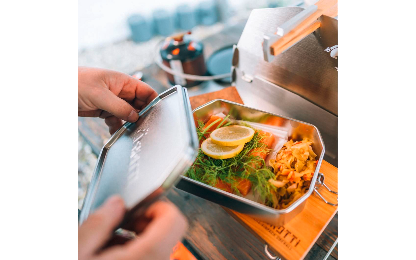 Main ouvrant une boîte à lunch métallique contenant deux rondelles de citron, des herbes vertes fines, des morceaux de nourriture orange et des légumes clairs râpés sur une table en bois