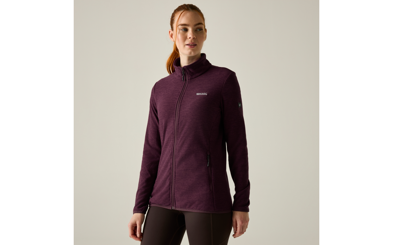 Persona con capelli rossi raccolti in una coda che indossa una giacca bordeaux con zip e piccolo logo sul petto e leggings scuri, in piedi davanti a uno sfondo chiaro, guarda leggermente verso destra