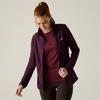 Donna con capelli rossi raccolti in una coda, guarda verso il basso e sorride, indossa una giacca con zip viola scuro aperta sopra una maglietta bordeaux e leggings neri su sfondo chiaro