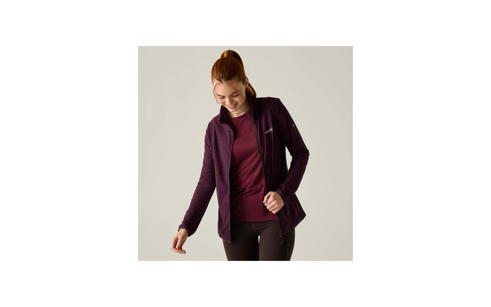 Donna con capelli rossi raccolti in una coda, guarda verso il basso e sorride, indossa una giacca con zip viola scuro aperta sopra una maglietta bordeaux e leggings neri su sfondo chiaro