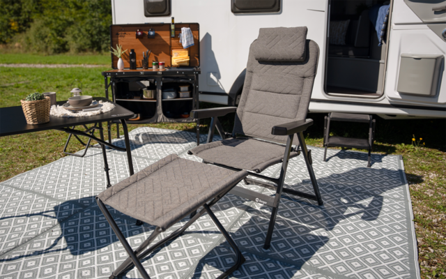 Gepolsterter grauer Liegestuhl mit Fußteil auf einem gemusterten Außen-Teppich vor einem offenen Wohnwagen; kleiner Tisch mit Tasse und Korb, Klapptreppe am Wohnwagen