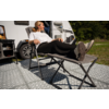 Person liegt auf einer zusammenklappbaren Campingliege vor einem Wohnmobil auf einem gemusterten Outdoor-Teppich, Bäume im Hintergrund